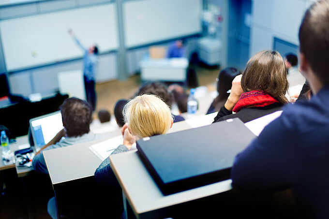 Speaker giving presentation in lecture hall at university. Participants listening to lecture and making notes.